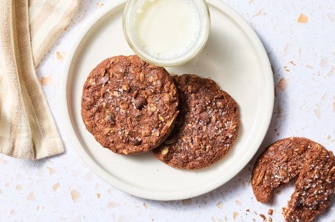 Two Chocolate Lactation Cookies on a plate with a glass of milk and another cookie on the counter next to the plate.