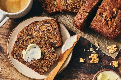 A slice of buttered Old-Fashioned Date Nut Bread on a plate atop a dark counter and with a latte and sliced loaf in the corner of the photo.