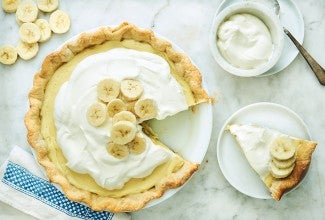 A Banana Cream Pie topped with fresh whipped cream and slices of banana with one slice of Banana Cream pie on a plate.