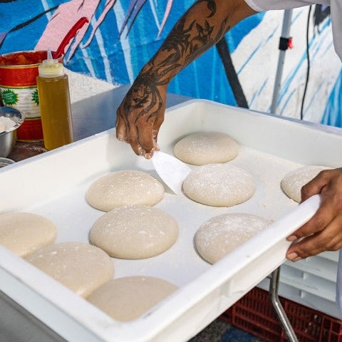 A person preparing raw balls of dough shown with them holding a white tub and lifting a dough ball with a spatula. - select to zoom