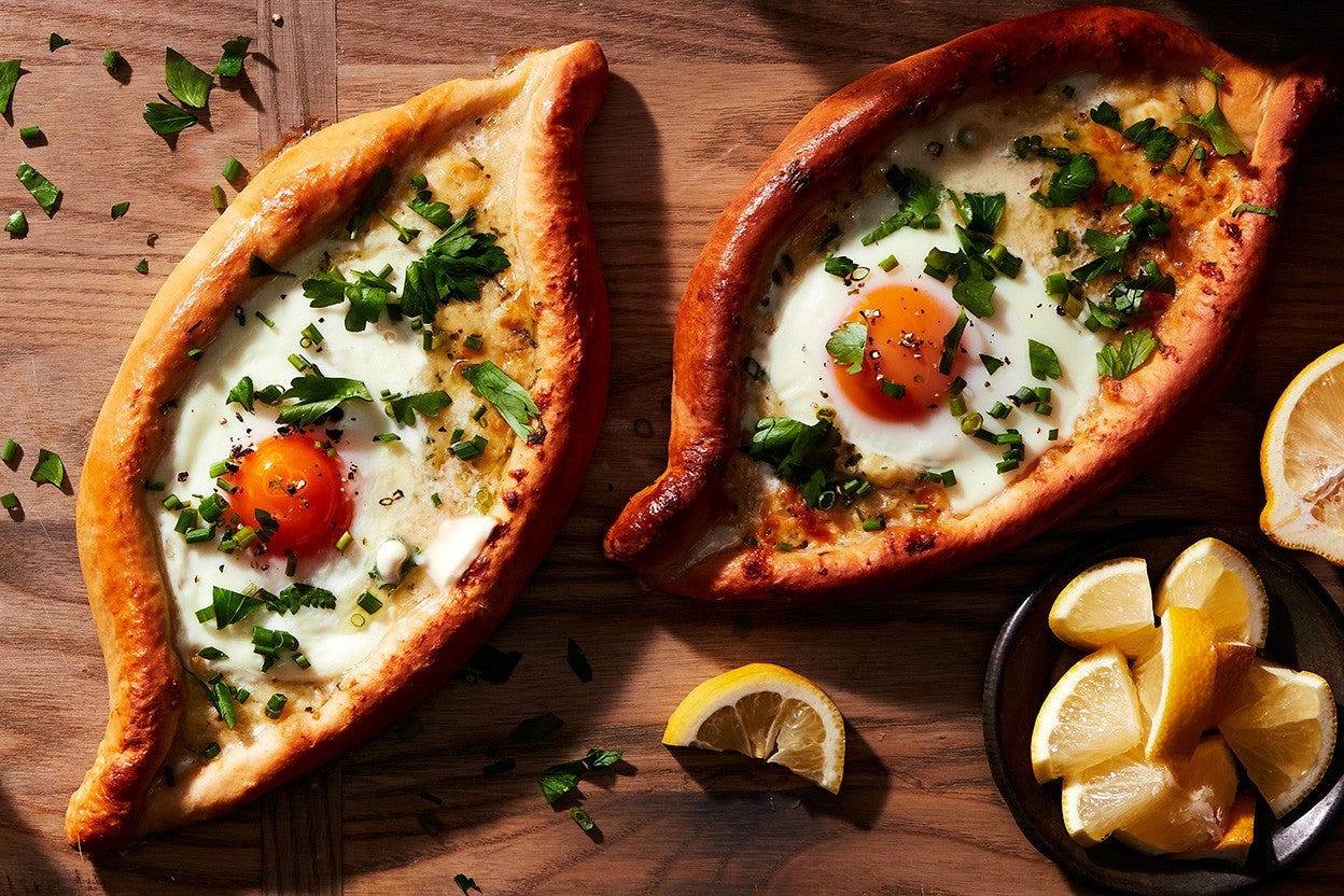 Two freshly baked Khachapuri (Georgian Cheese Breads) topped with soft-baked eggs and fresh herbs