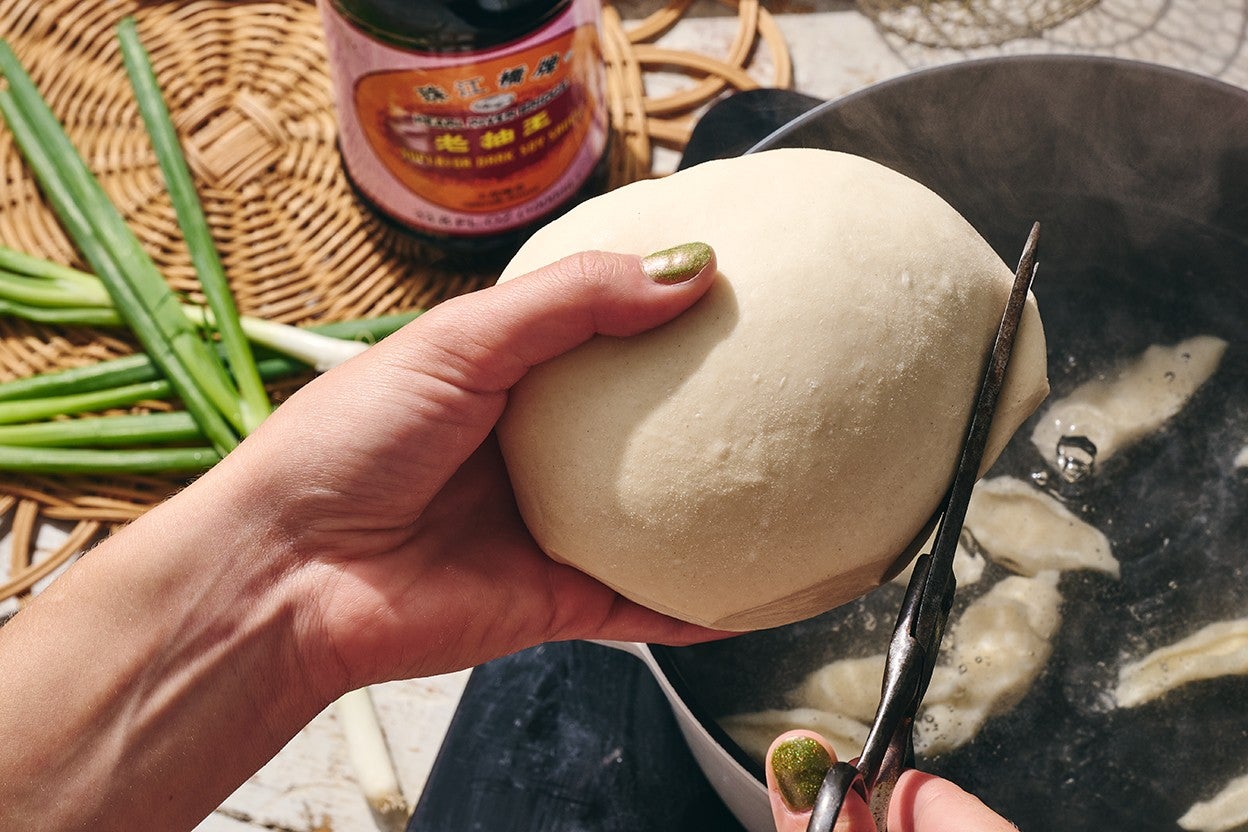 A hand holding a ball of noodle dough and cutting noodles into a pot of boiling water.