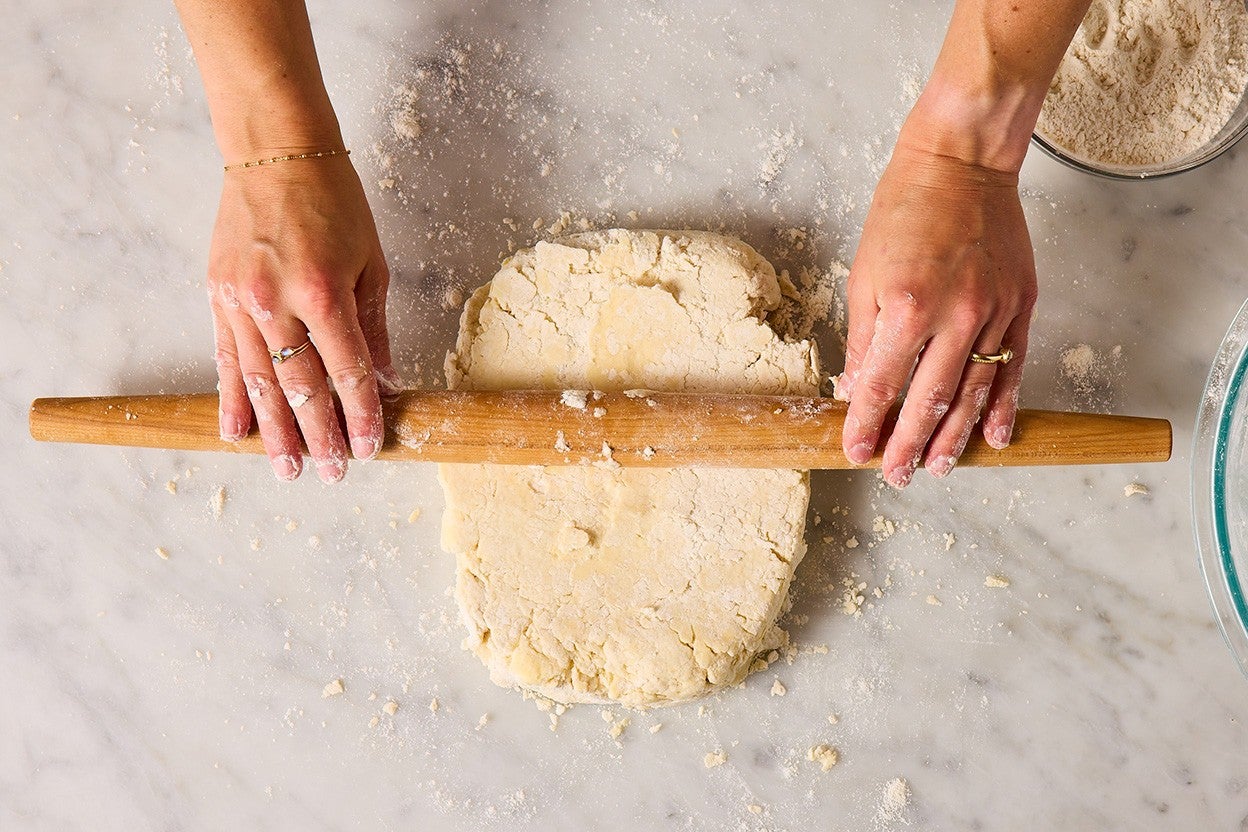 Two hands holding either side of a long rolling pin to press and roll biscuit dough on a marbled countertop.