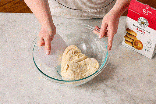 Overhead view of shaggy bagel dough being mixed in a bowl with a box of King Arthur Baking Chewy Bagel Mix in the bottom right corner of the frame.