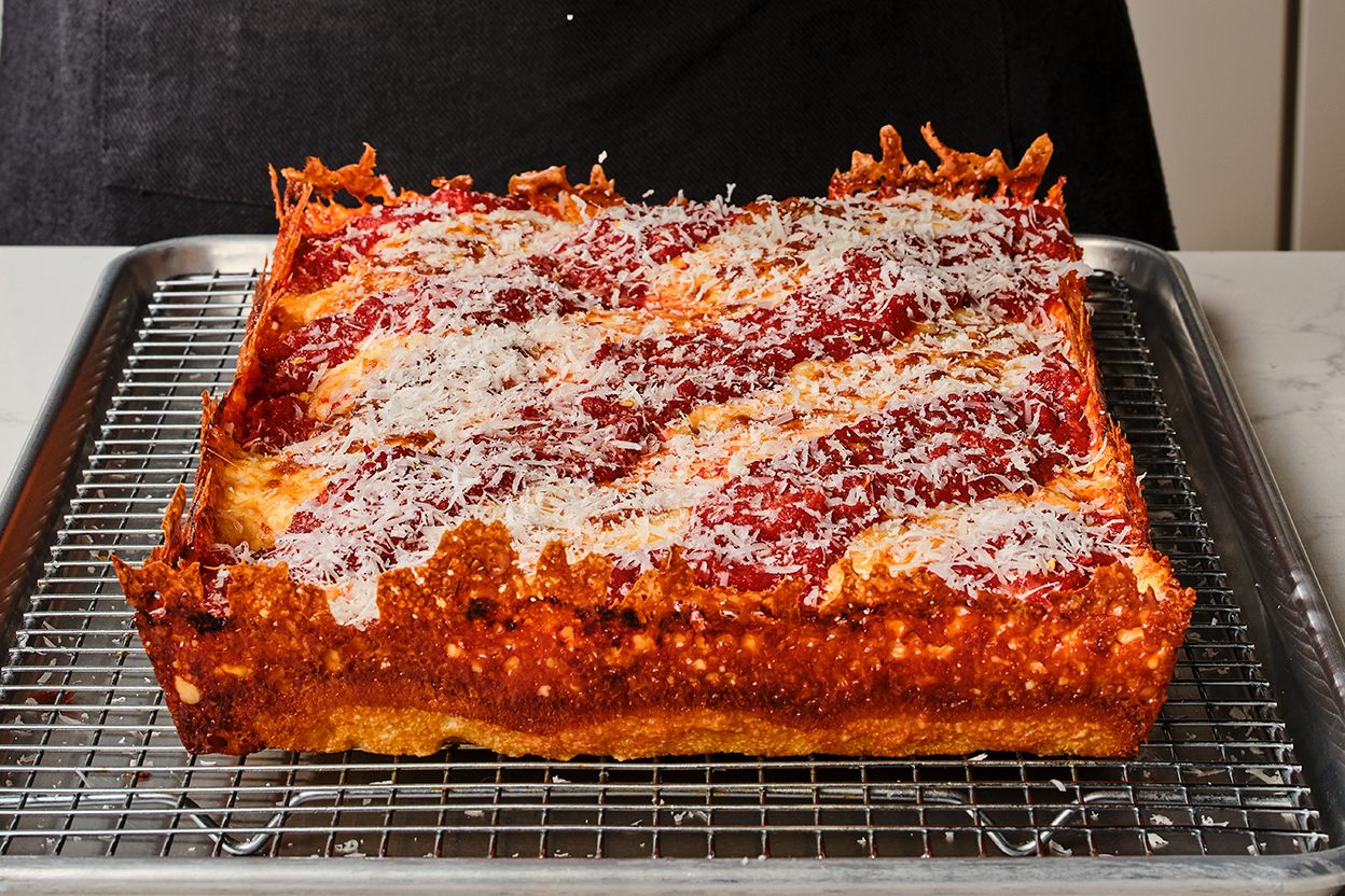Detroit pizza on a baking rack showing the cooked pizza edges with crispy cheese and dough and a thick overall pizza.
