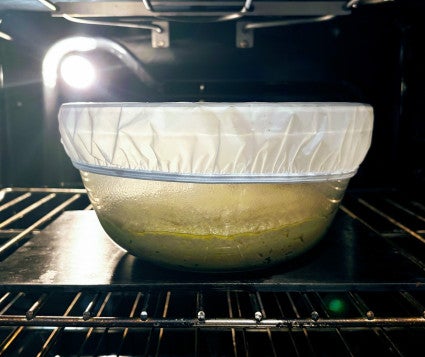 Yeast dough risinng in clear glass bowl (for visibility), covered with a fitted plastic bowl cover, on middle rack of oven with light turned on.