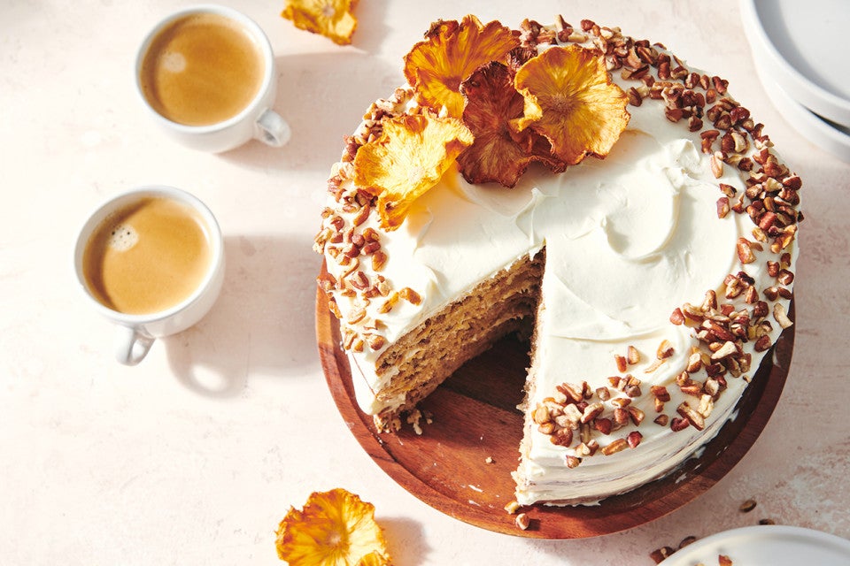 An overhead photo of a Hummingbird Cake with pineapple cream cheese frosting. A slice has been taken out of the cake and there are toasted nuts and a slice of dried pineapple on top of the cake. 
