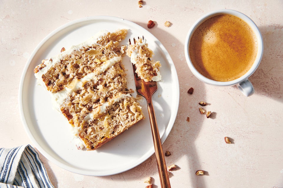 An overhead photo of a slice of Hummingbird Cake on a plate with a fork resting on the edge. You can see the tender cake layers with cream cheese pineapple frosting sandwiched between them. 