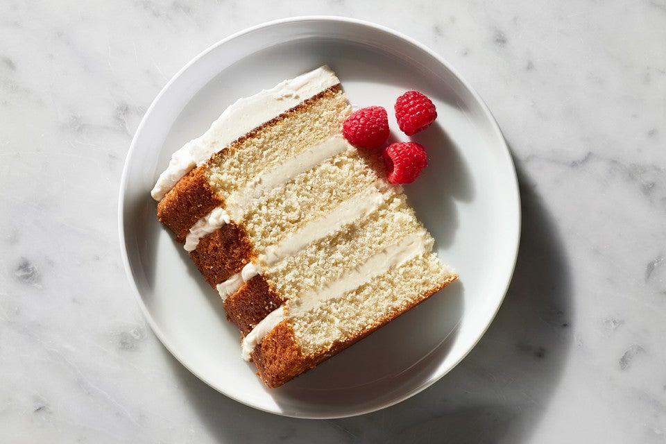A slice of a four-layer Tender White Cake filled with vanilla buttercream and garnished with raspberries on a plate