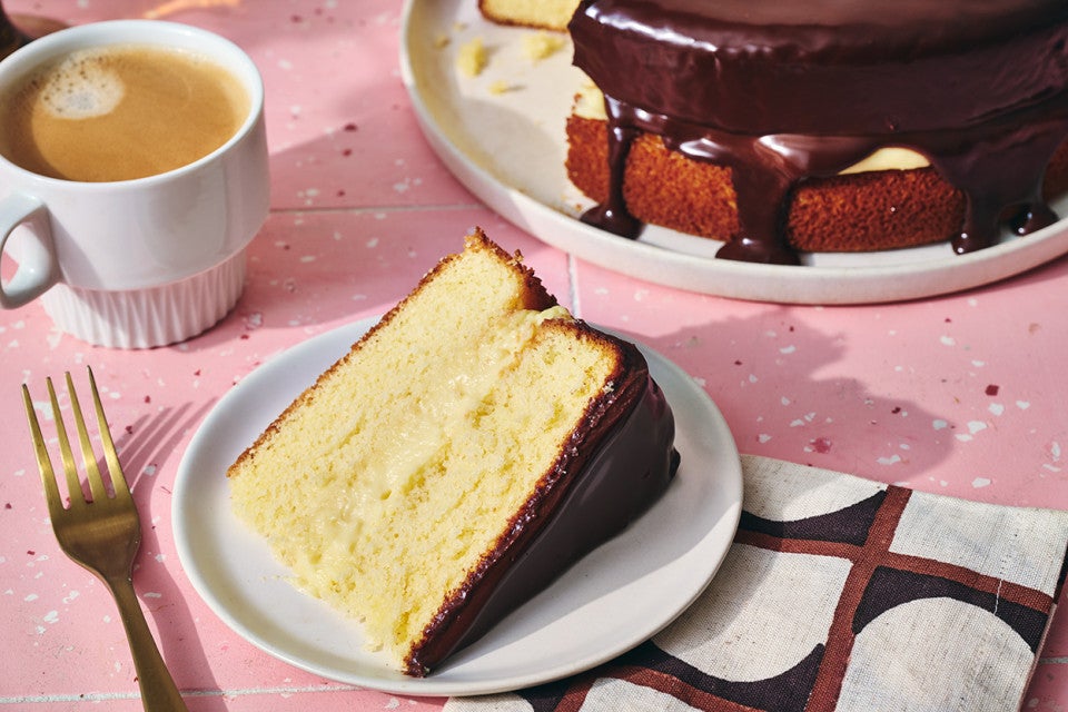 A slice of Boston Cream Pie on a plate, filled with vanilla pastry cream and topped with chocolate ganache.