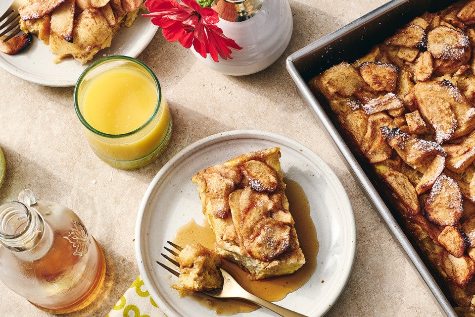A slice of Apple French Toast Bake on a light colored plate with syrup spilling out beneath the french toast. The scene also included a cup of orange juice, syrup in a jar, and shows the corner of the baked french toast in a baking pan.