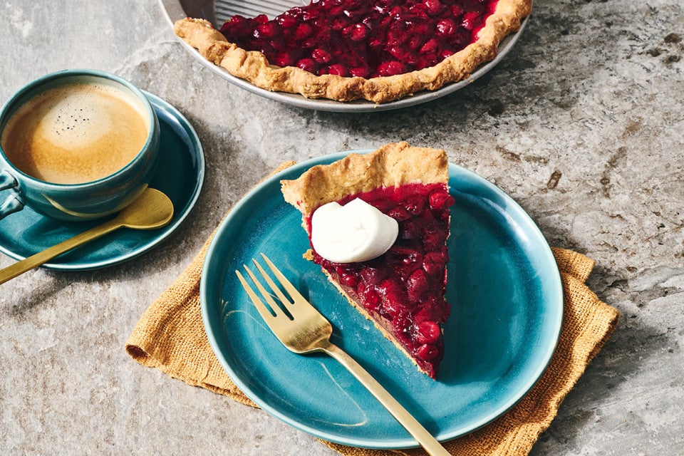 A slice of Cranberry-Fudge Pie topped with a dollop of whipped cream, a cup of coffee sits on a plate off to the side.