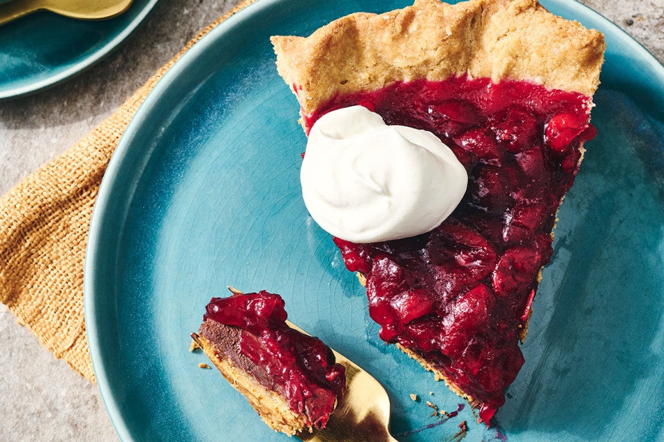 Cranberry-Fudge Pie seen from above with dark and glossy cranberries, a dollop of whipped cream, and a beautifully browned crust. A bite on a fork show the fudgy bottom of the pie.
