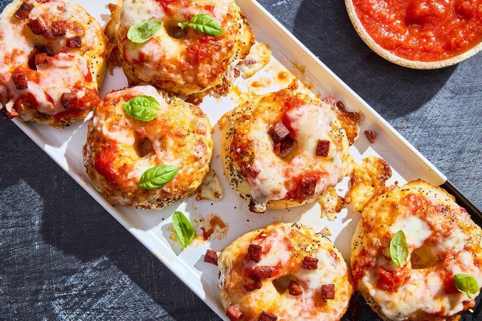 Delicious mini pizza bagels arranged on a plate showing melty cheese, pepperoni, and basil with a bowl of marinara in the top of the photo.