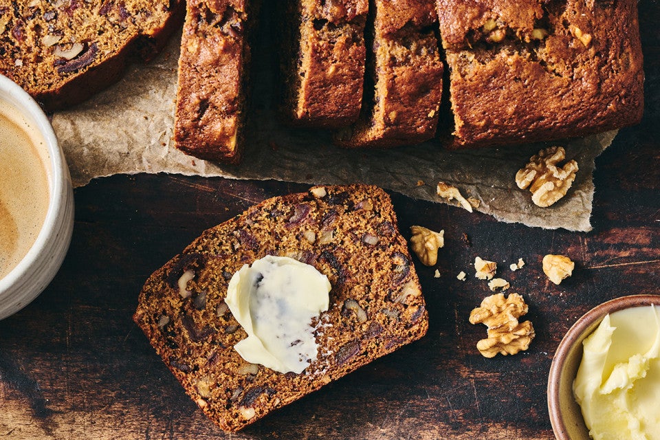 A slice of buttered Old-Fashioned Date Nut Bread on a plate atop a dark counter and with a latte and sliced loaf in the corner of the photo.