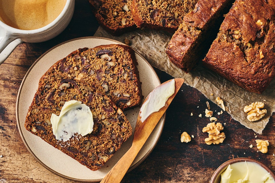 A slice of buttered Old-Fashioned Date Nut Bread on a plate atop a dark counter and with a latte and sliced loaf in the corner of the photo.