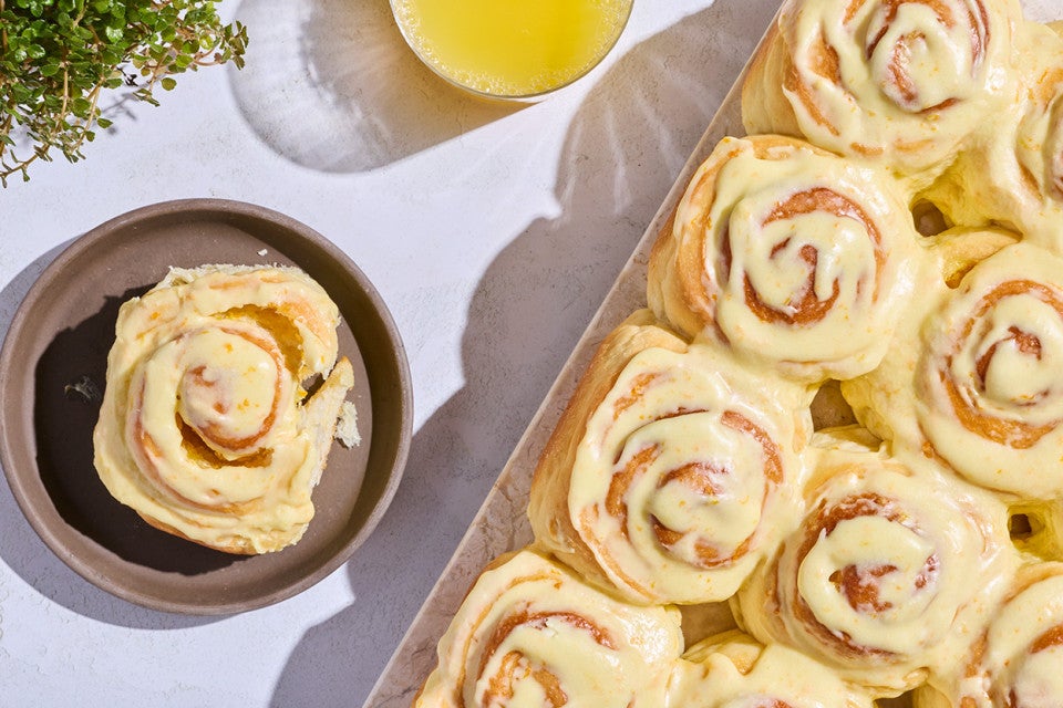 Fluffy Frosted Orange Rolls seen from above on a sheet tray with a single roll on a dark plate.