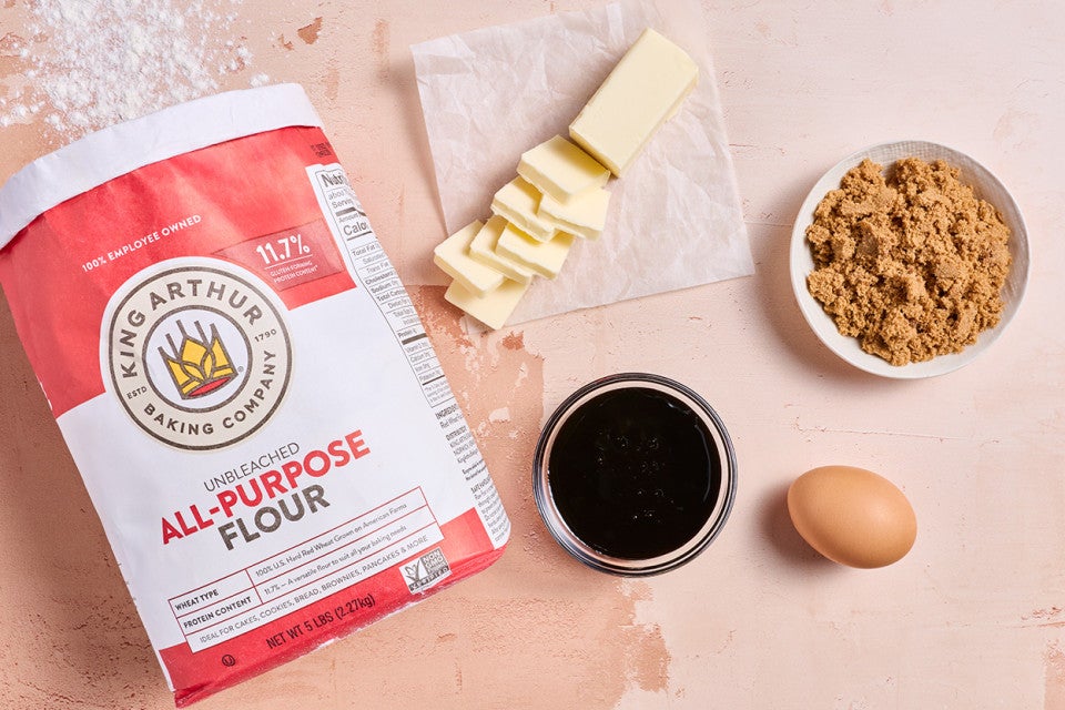 Ingredients to make Soft Brown Sugar Cookies laid out on a warm countertop showing butter, brown sugar, an egg, vanilla, and all-purpose flour.