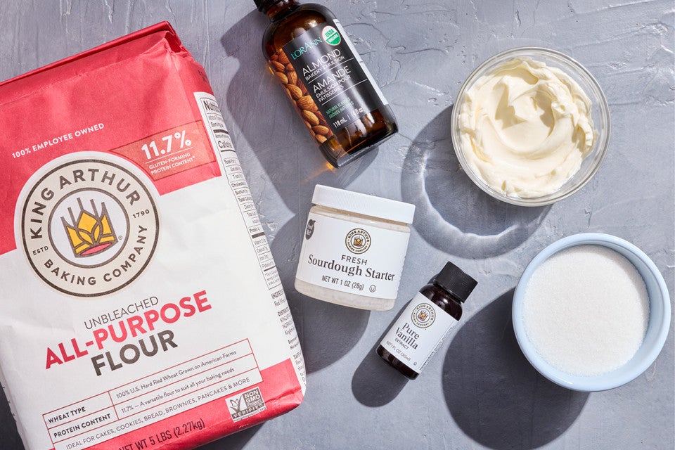 Ingredients to make Sourdough Sugar Cookies - flour, sourdough starter, almond extract, vanilla extract , sugar, and cream cheese - shown on a counter from above.