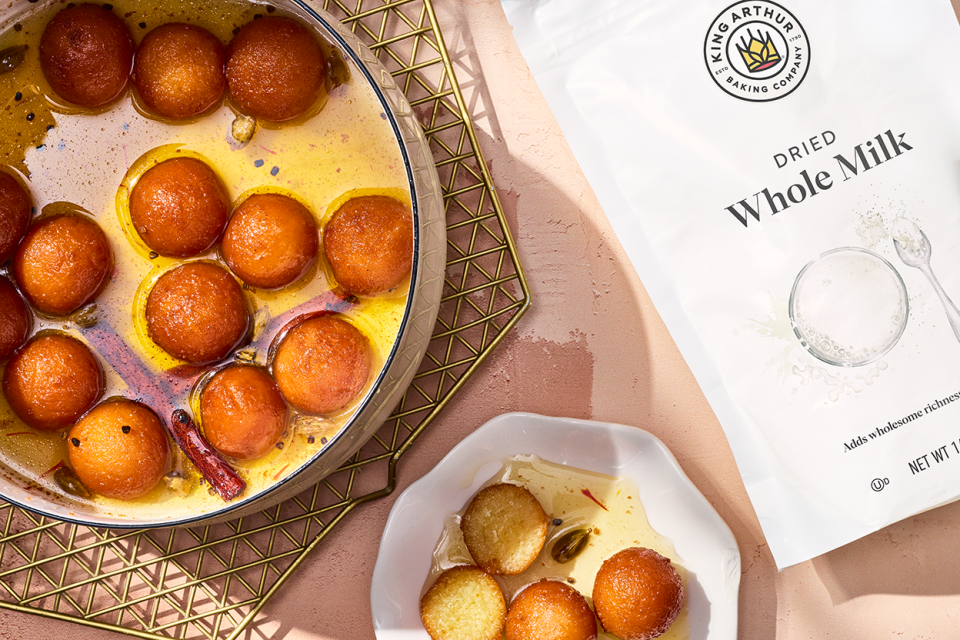 Gulab Jamun in a pot and on a plate with King Arthur Dry Whole Milk Powder in a bag next to the desert.