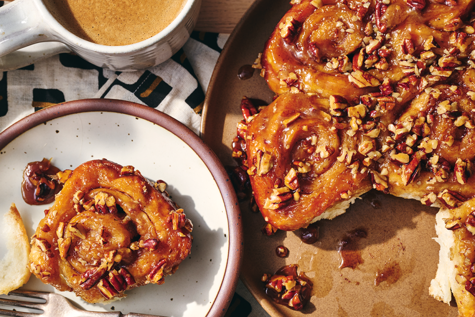 A single sticky bun on a plate adjacent to freshly baked buns.