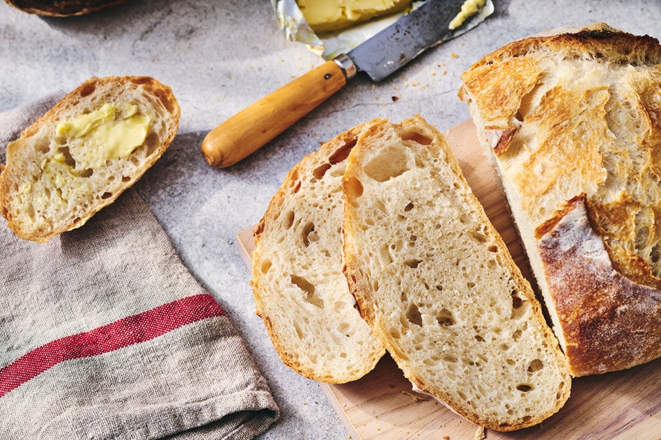 Fresh baked loaf of No-Knead Crusty White Bread on a table with jam and butter and slices on a cutting board.