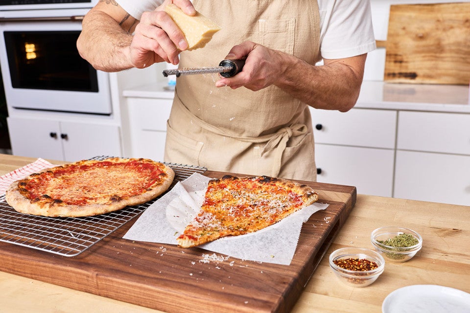 Martin grating cheese over a slice of New York-Style Pizza on a counter.