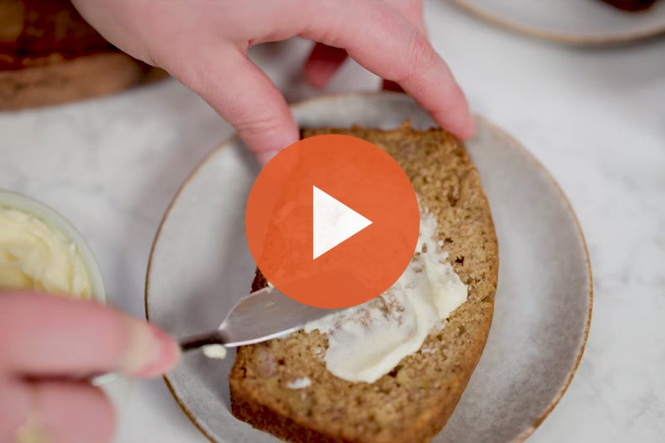 Spreading butter on banana bread with a play button to watch the video.