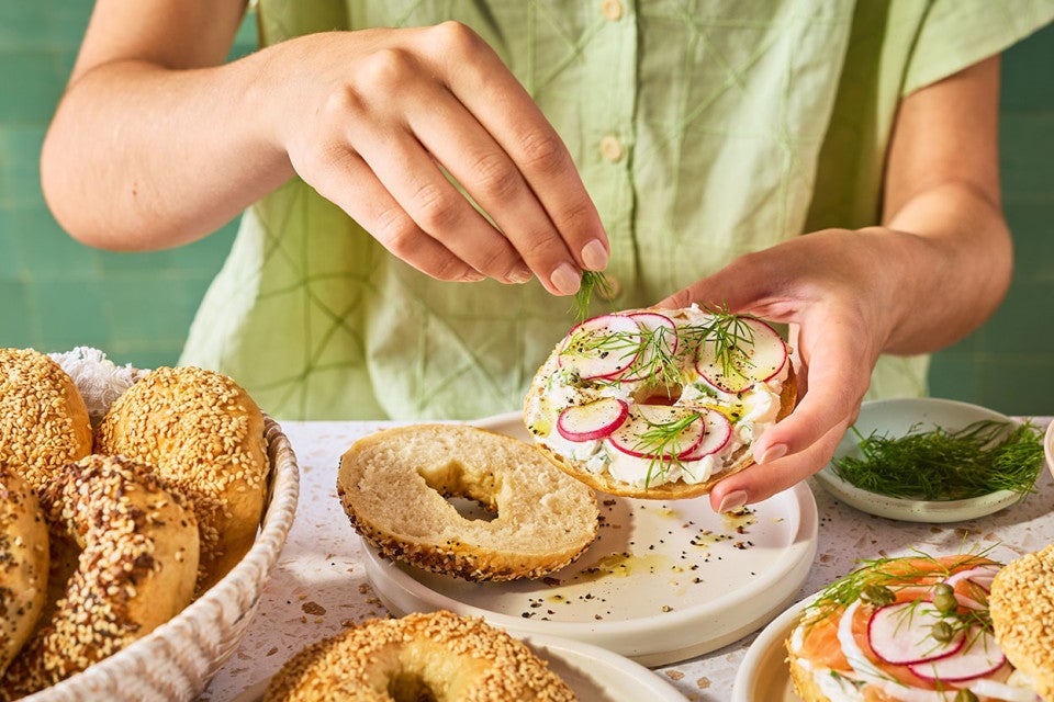Someone sprinkling seasoning on a dressed bagel.