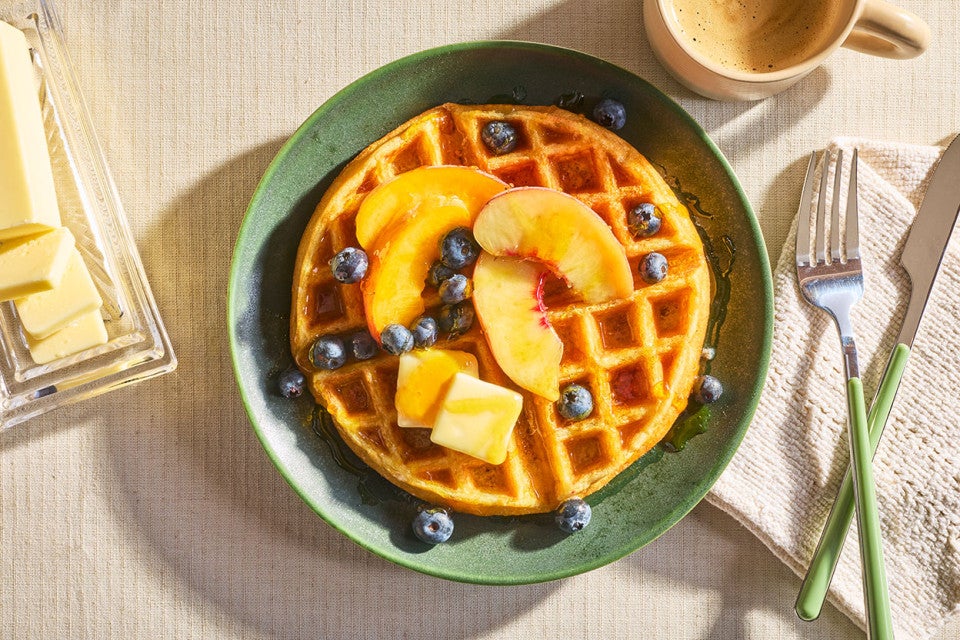 A Classic Buttermilk Waffle on a green plate and topped with butter, syrup, peaches, and blueberries.