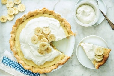 A Banana Cream Pie topped with fresh whipped cream and slices of banana with one slice of Banana Cream pie on a plate.