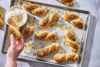 A hand dipping an Everything Bagel Twist into cream cheese along with other bagel twists spread out on a sheet tray and parchment paper.