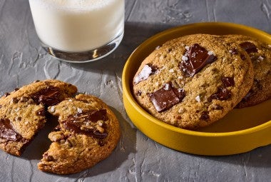 Beautifully baked High-Altitude Chocolate Chip Cookies shown on a plate and counter with a glass of milk in the background.