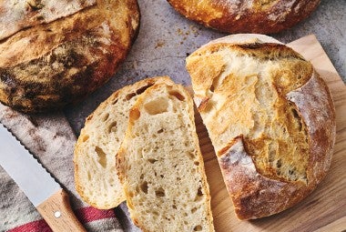 Fresh baked loaves of No-Knead Crusty White Bread on a table with jam and butter and slices on a cutting board.