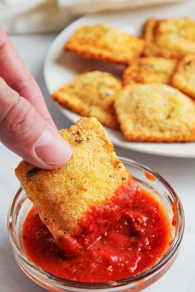 Hand dipping fried ravioli in a bowl of marinara sauce with fried ravioli in the background on a white plate