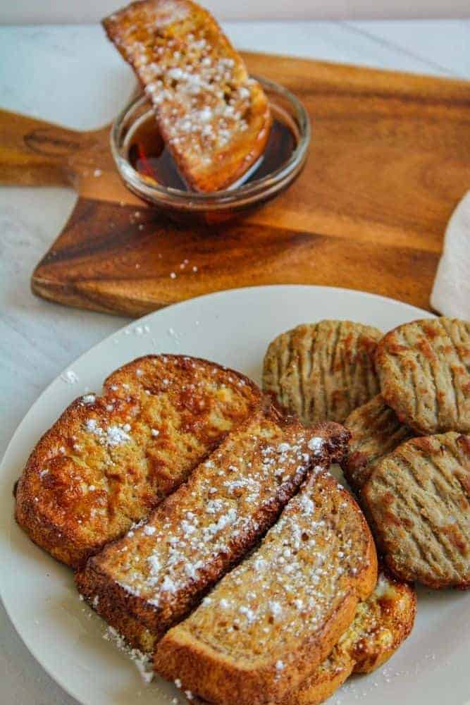 Plate of Air Fried French Toast with sausage on a plate and one stick being dipped into maple syrup in background