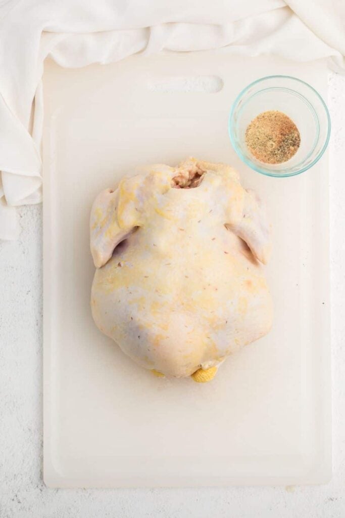 Raw whole chicken on a cutting board with seasoning in a bowl