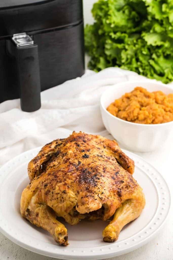 Full cooked whole chicken on a white seving plate with mashed sweet potatoes in a whie bowl, air fryer, and head of lettuce behind it