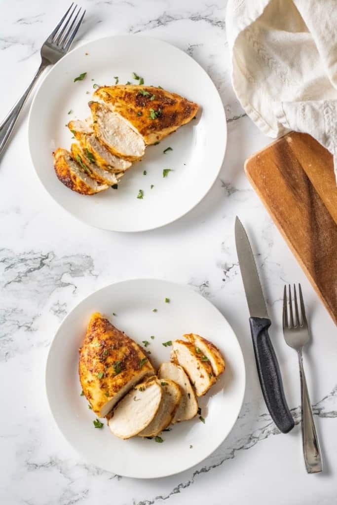 Overheard view of two white plates of frozen chicken cooked in an air fryer sliced next to a fork and knife