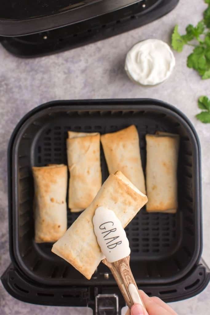 Cooked frozen burritos in air fryer with tongs grabbing one burrito