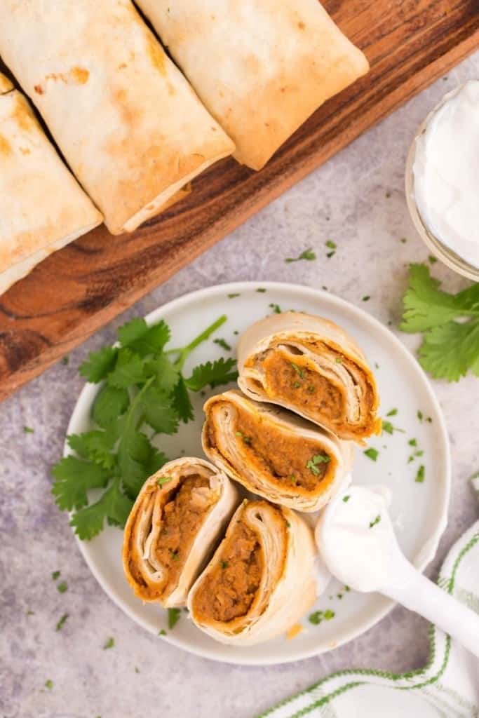Burritos cut in half on a serving plate with sour cream and full burritos in background