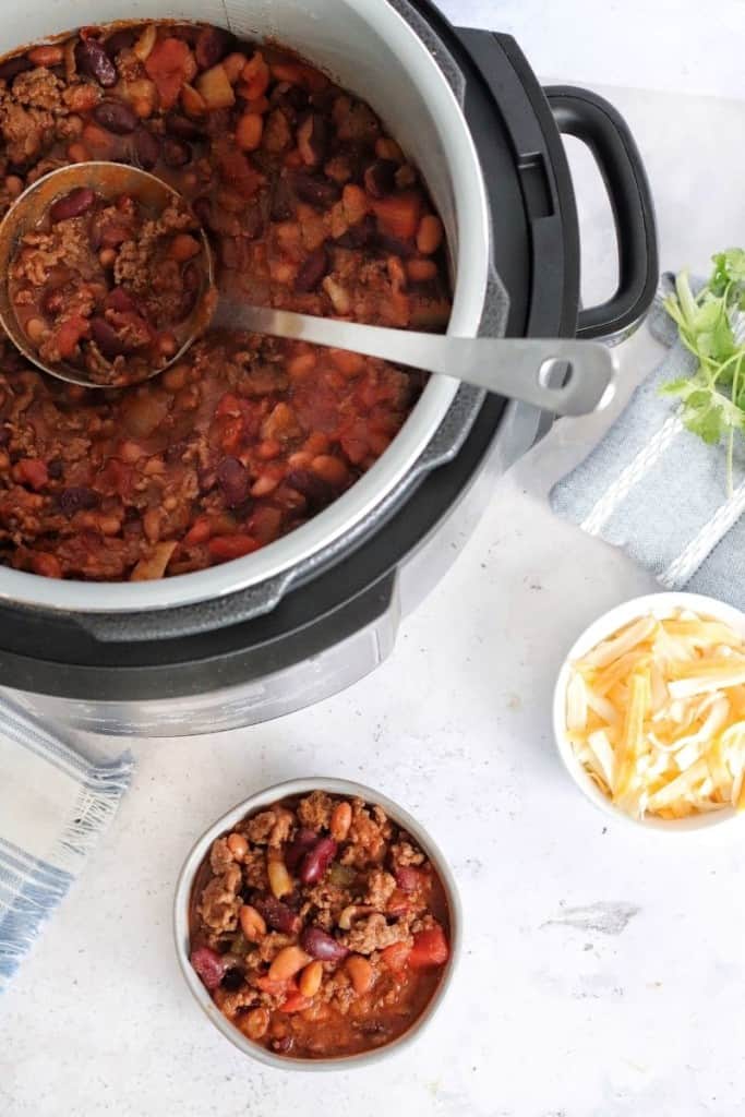 Chili inside the Ninja Foodi with a serving ladle inside and a bowl of chili and cheese to the side