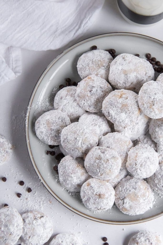 Snowball cookies on a round grey plate with snowball cookies and chocolate chips to the side of it