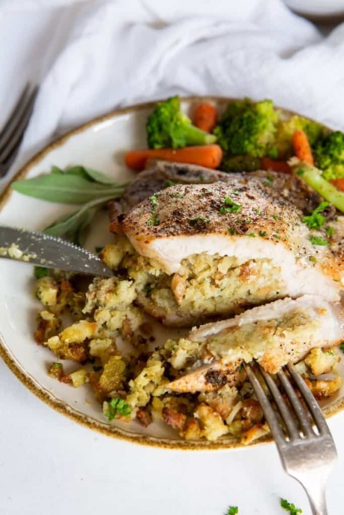 Sttuffed pork chop cut in half with stuffing showing on a white plate served with veggies (broccoli and carrots)