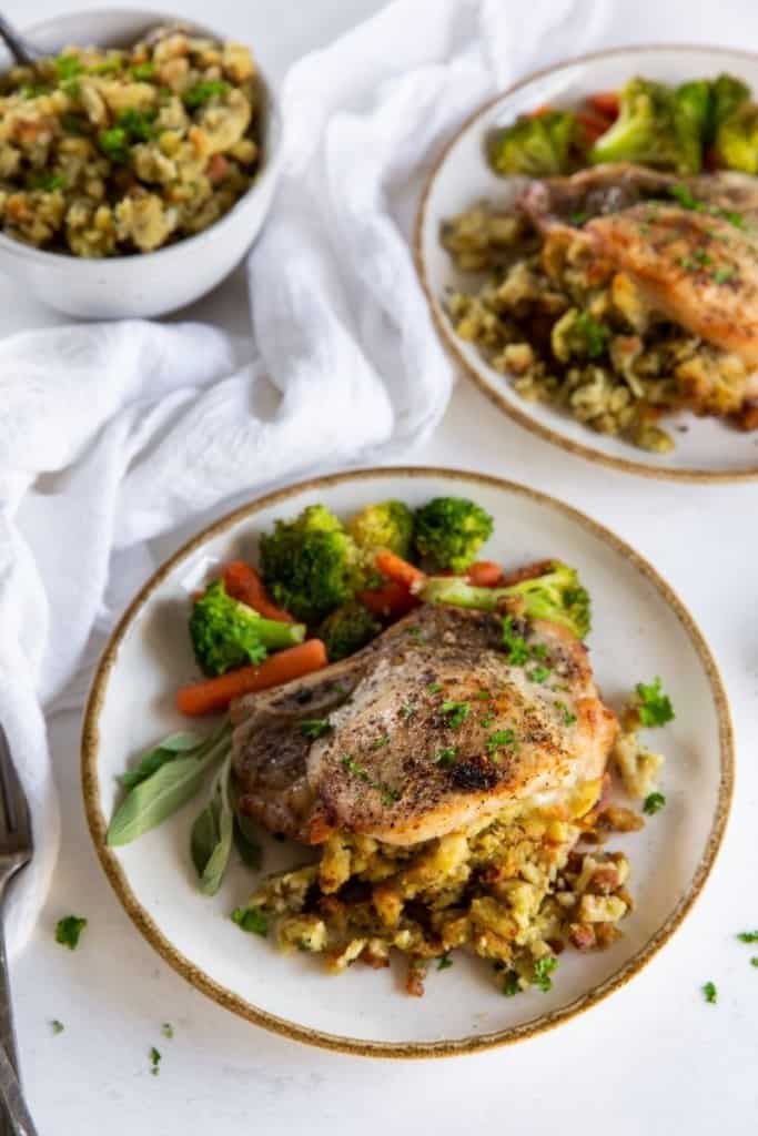 Pork Chops stuffed with stovetop dressing on a white plate with broccoli and carrots with an extra bowl of dressing in background