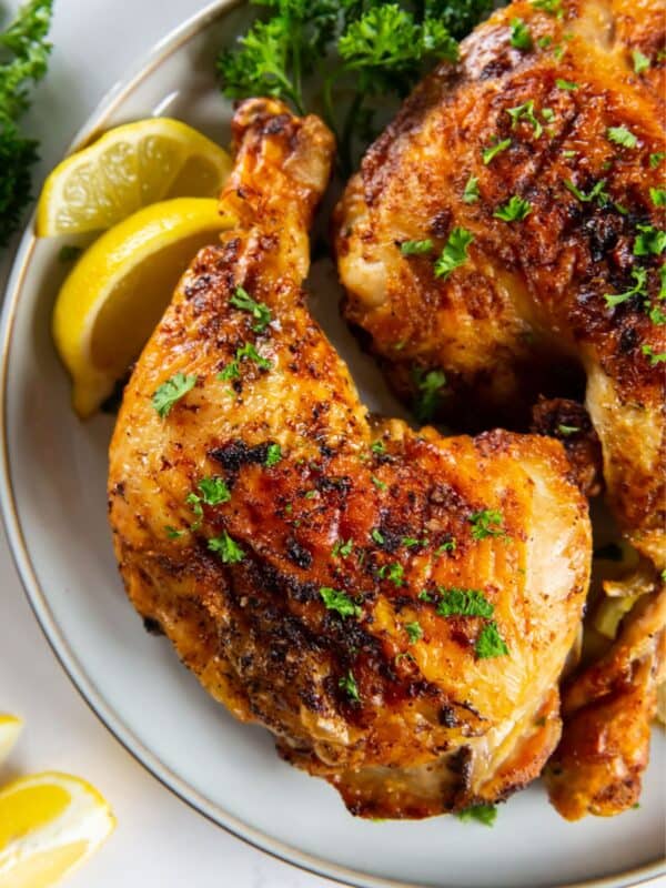 Close up of air fried chicken leg quarters on a white plate served next to lemon slices.