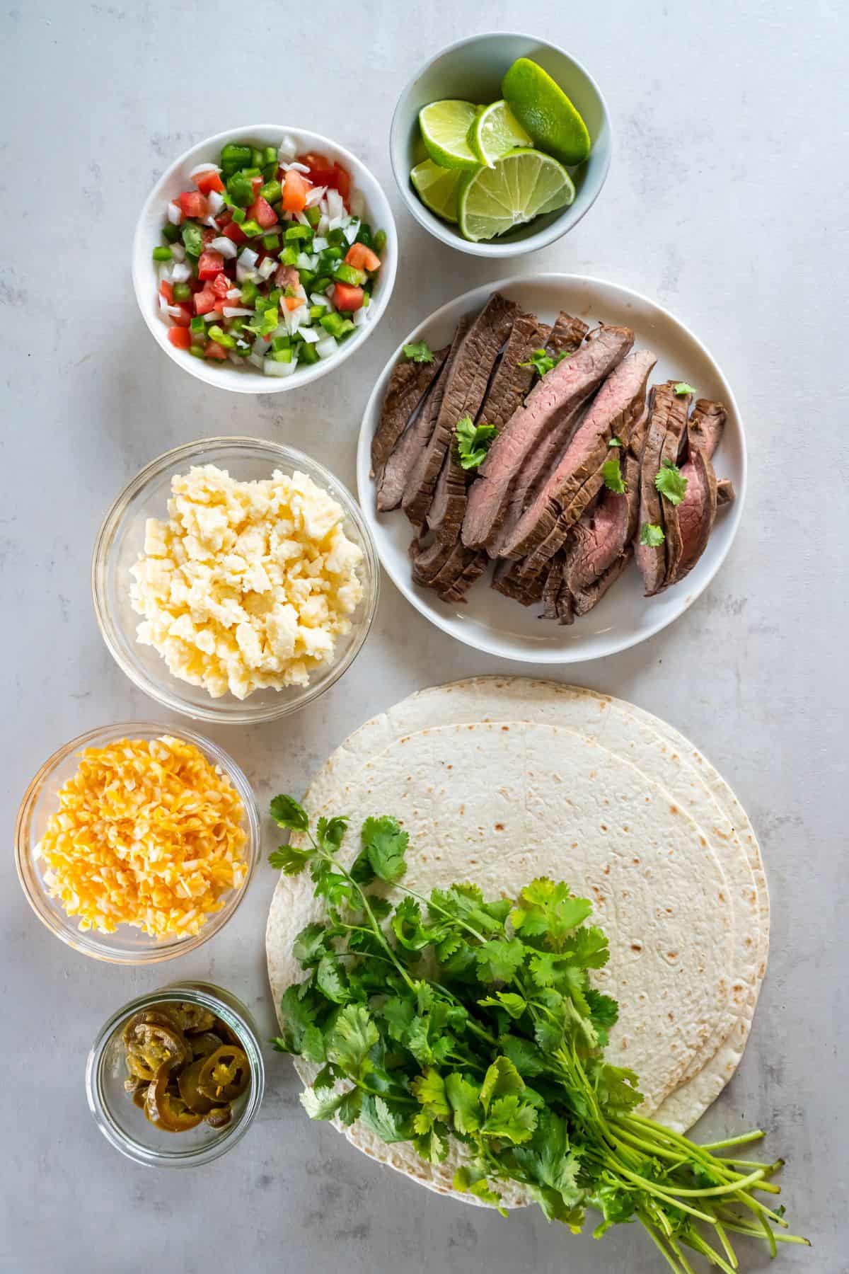 Ingredients needed to make carne asada quesadillas: tortillas, quesadilla cheese, pico de gallo, jalapeno slices, cheddar cheese, and cilantro.
