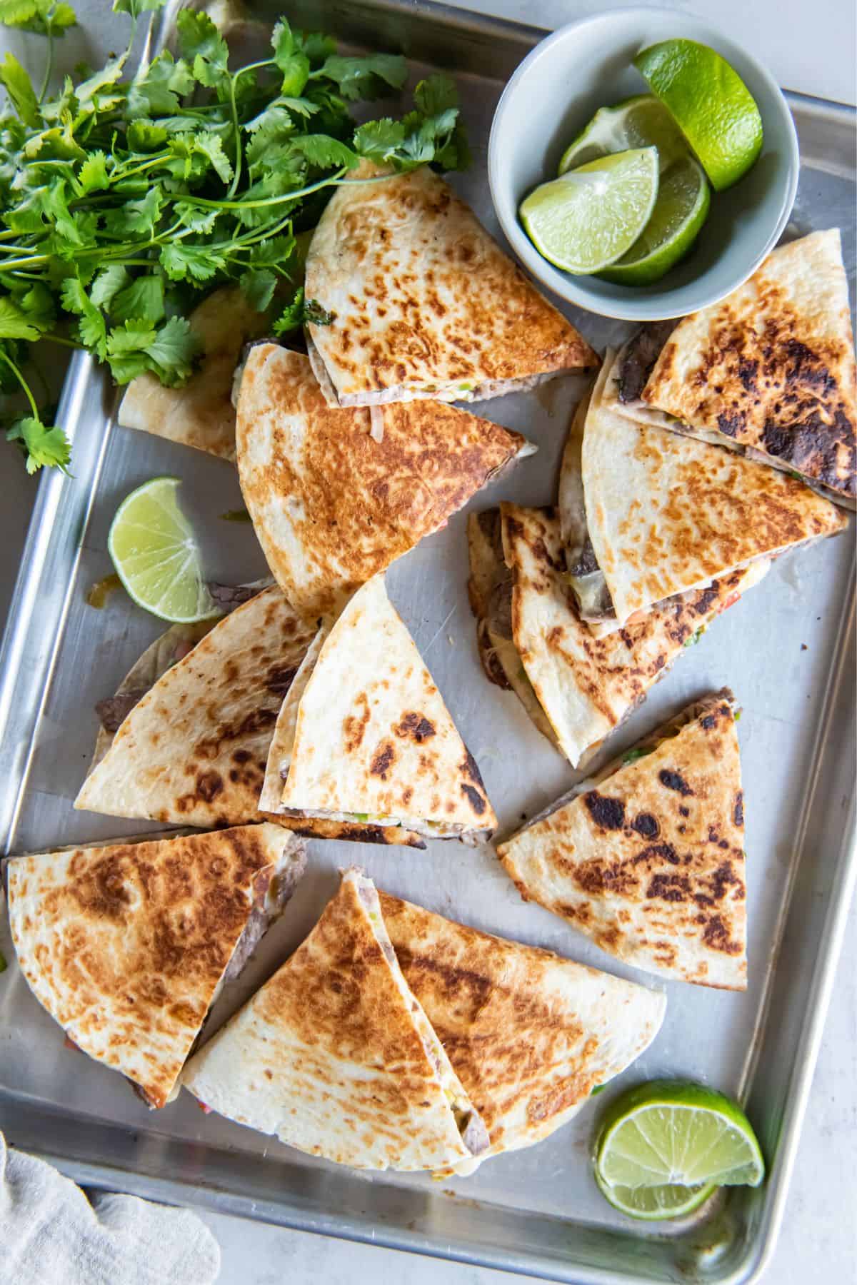 Steak quesadillas cut into slices on a baking sheet serving tray with limes.