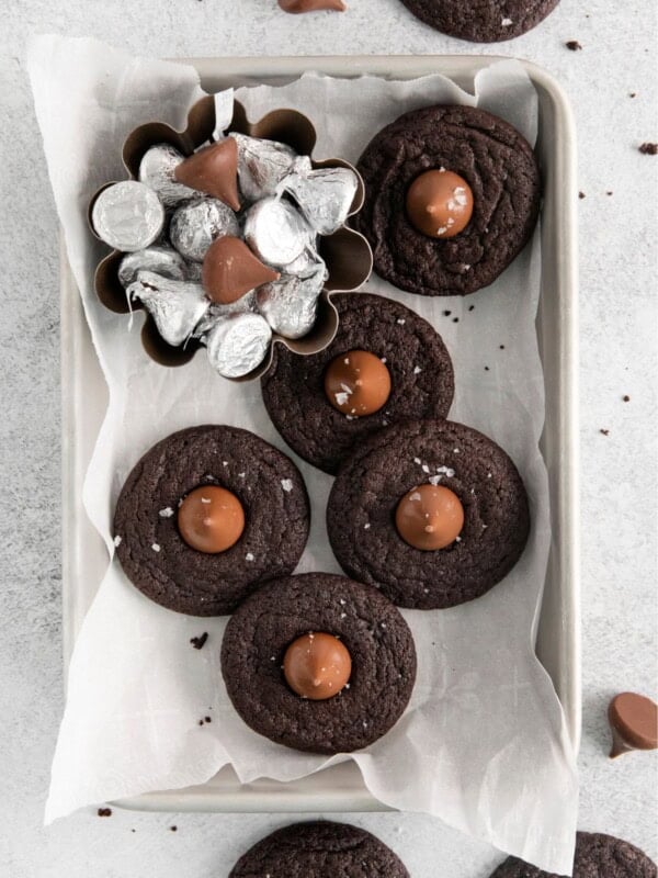 Overhead view of chocolate kiss cookies on parchment paper in a storage tin and a little bowl of Hershey kisses on the side.