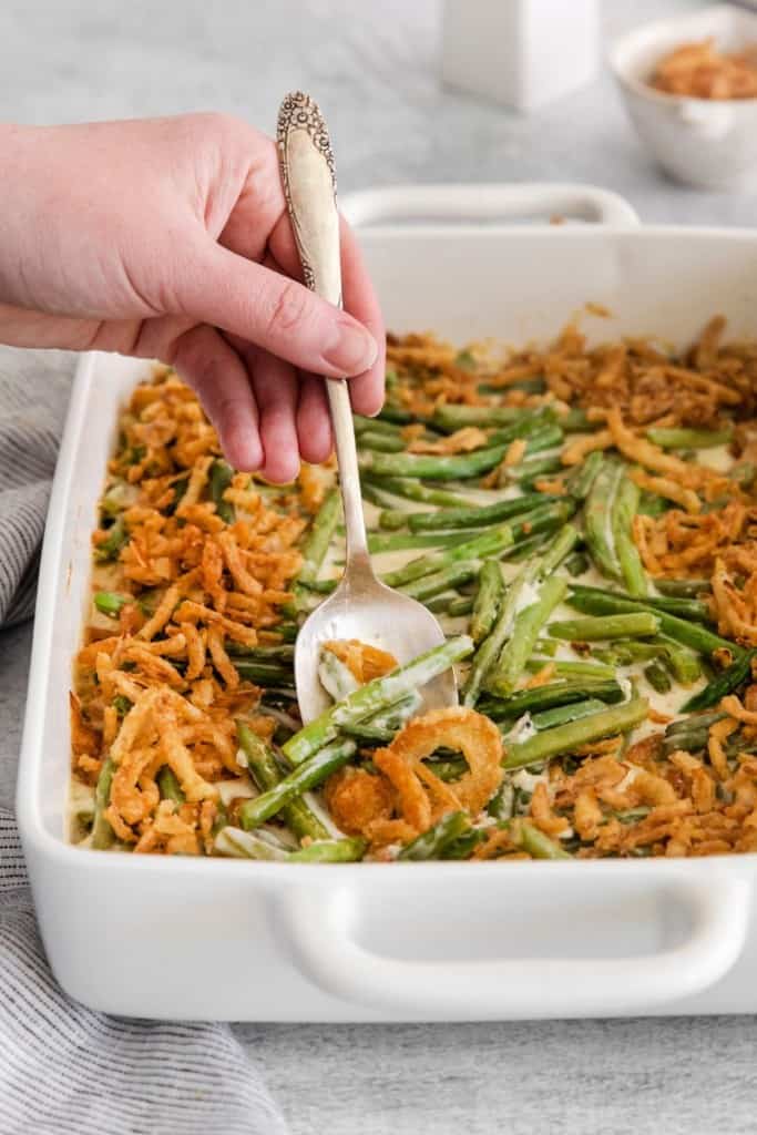A spoon taking out some homemade green bean casserole with fried onions on top from a baking dish.